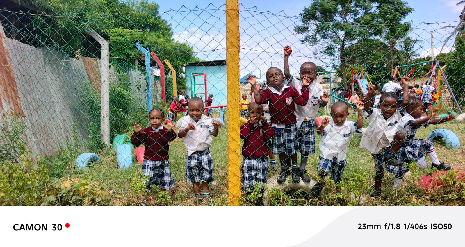 A different group of kindergarten pupils in maroon and white uniforms posing at the playground fence